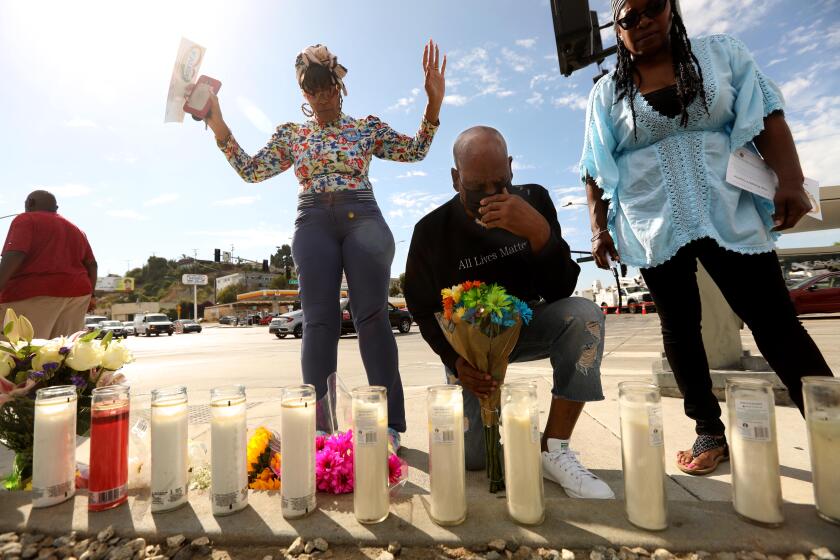 LOS ANGELES, CA - AUGUST 5, 2022 - - Carmen Dorsey, from left, and London Carter pray as Vera Jones lights a candle at a makeshift memorial across the street from where a fiery multi-car crash left six dead, including a pregnant woman, and injured others in Windsor Hills in Los Angeles on August 5, 2022. The accident happened on Thursday, August 4, 2022. (Genaro Molina / Los Angeles Times)