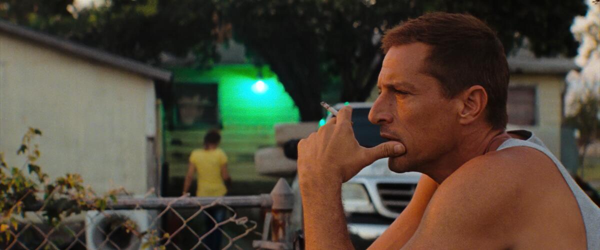 A man sitting outdoors by a chain-link fence smokes a cigarette and looks contemplative.