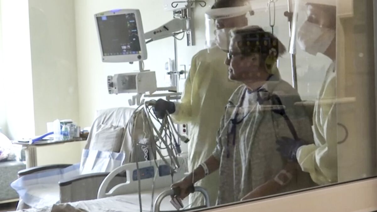 A person in hospital gown using a walker is flanked by two people wearing masks, face shields and gloves in a hospital room
