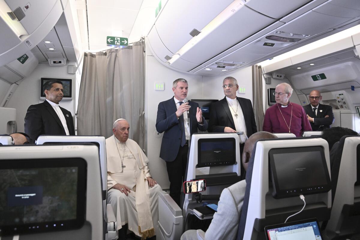 Five men stand and one man in a cassock sits in an airplane while facing other passengers.