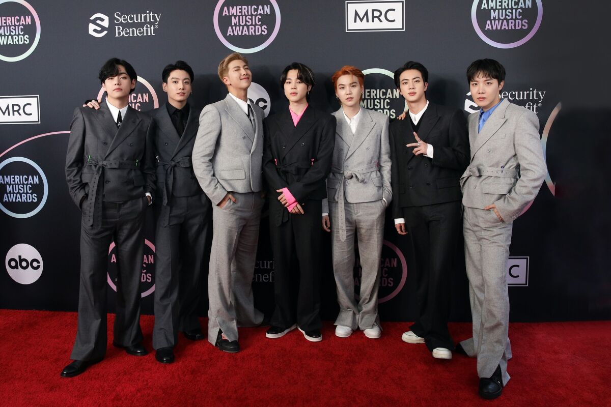 Seven men posing on a red carpet in black and gray suits