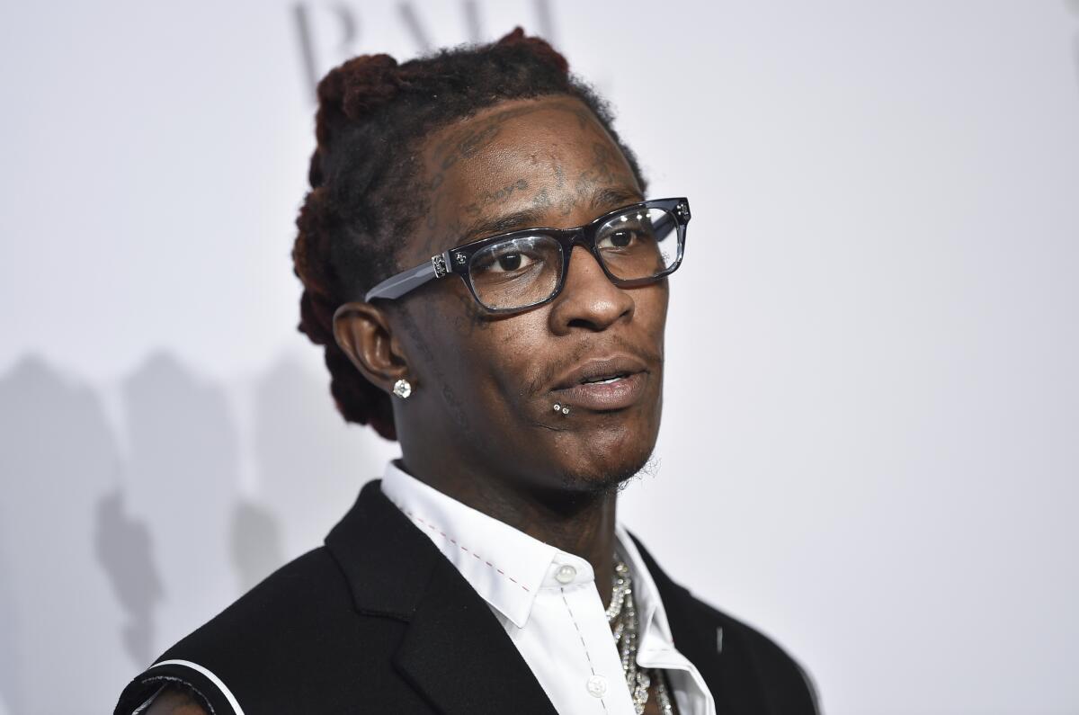 Young Thug posing, wearing thick-framed glasses and a black vest and white button-up shirt