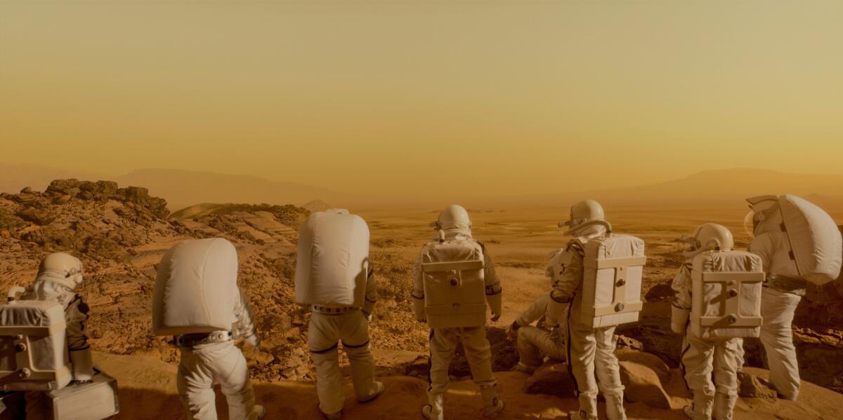 Seven people in white spacesuits are seen from behind looking out over a desert landscape with an orange haze.