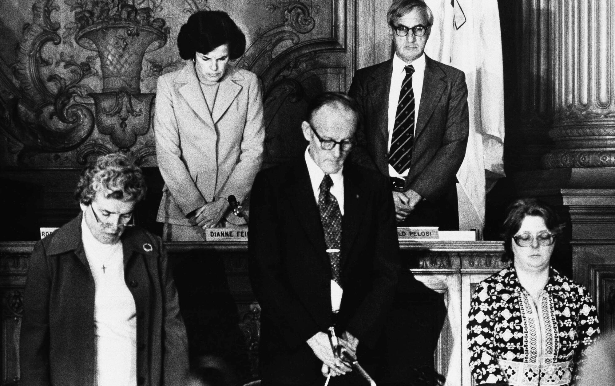 A black-and-white image  of five people, including then Mayor Dianne Feinstein, top left, bowing their heads
