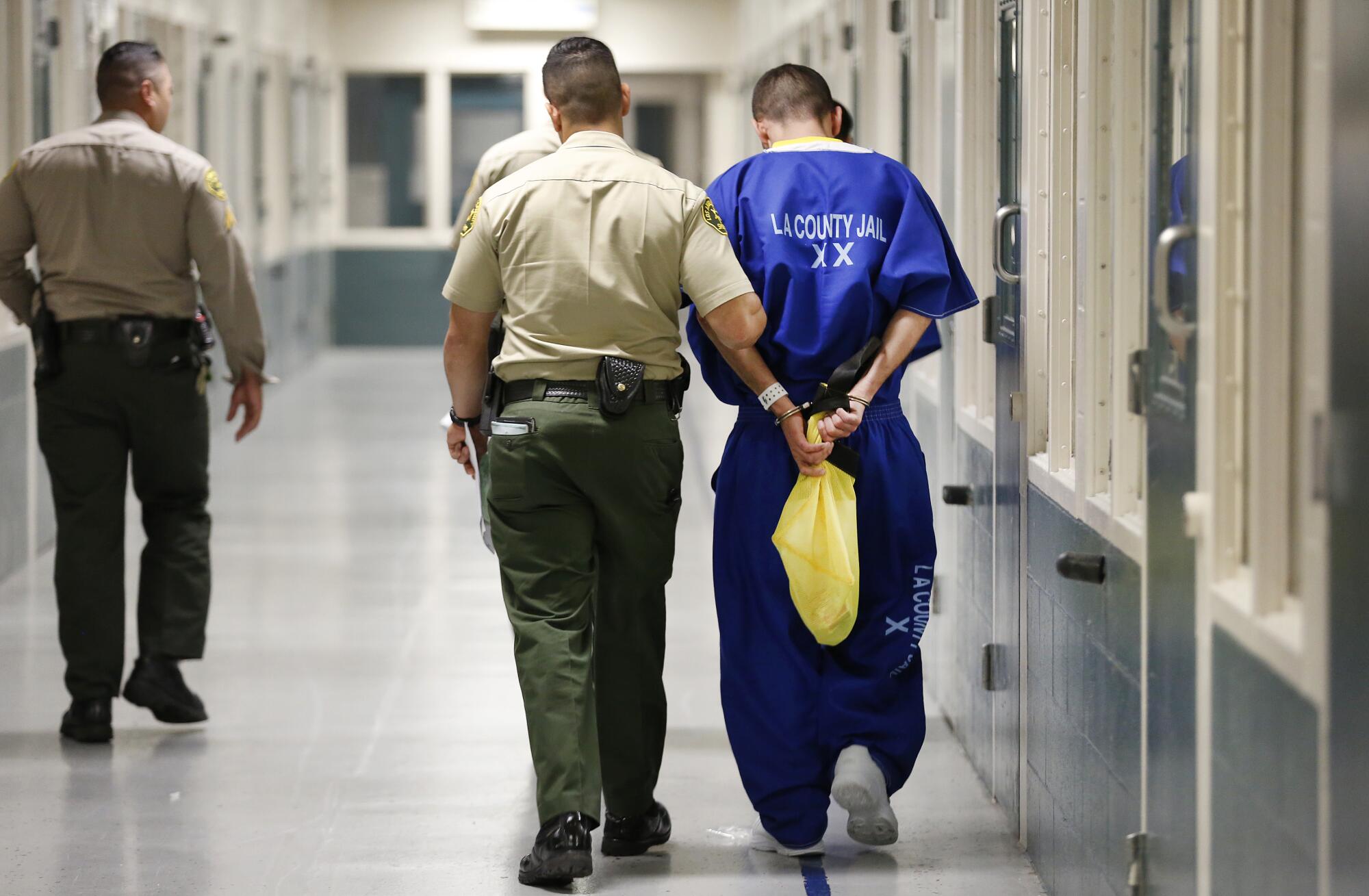 A uniformed deputy escorting a handcuffed detainee in a blue jumpsuit through a hallway