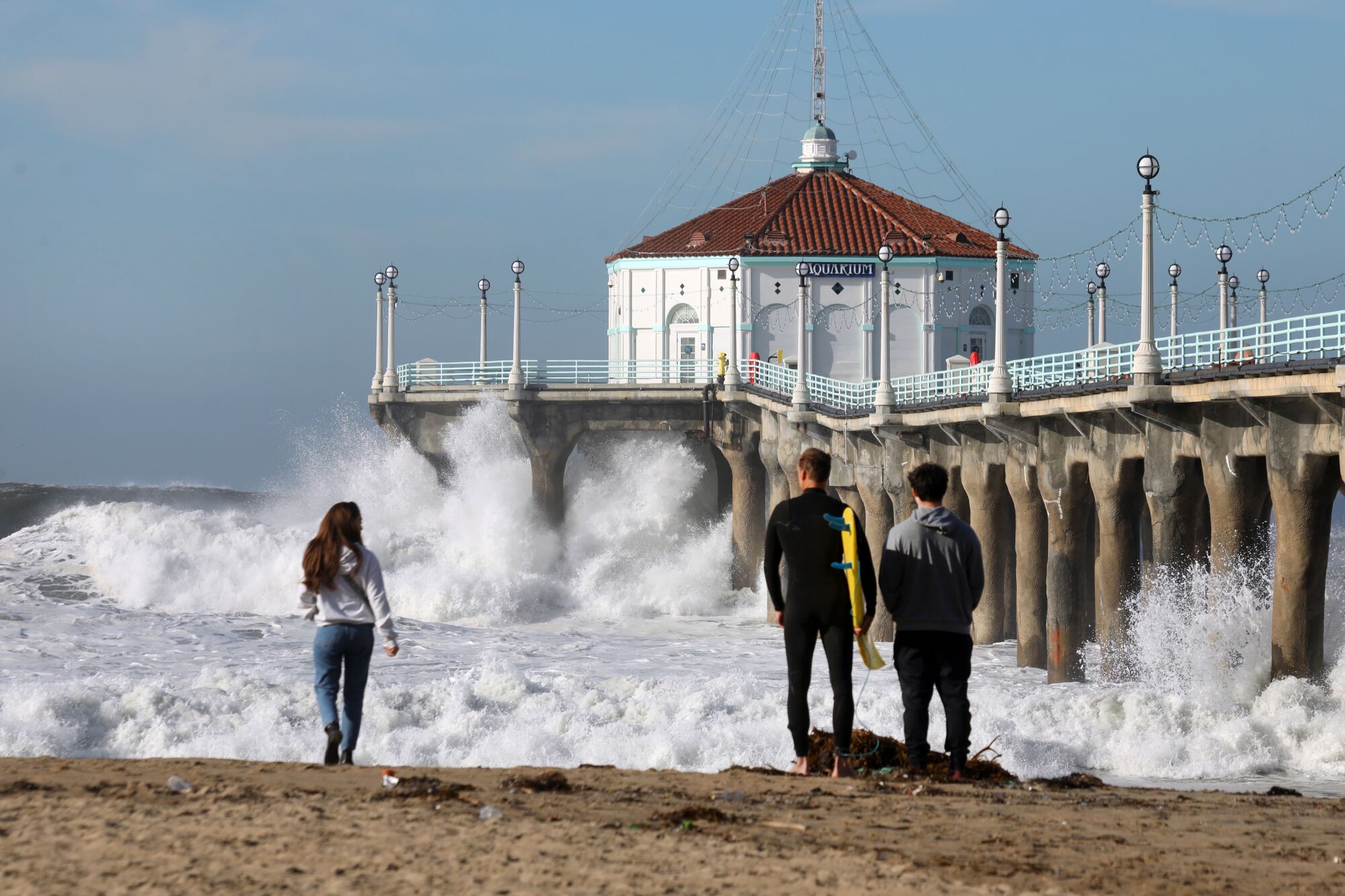 Three people, the middle one holding a surfboard, stand on the beach, a pier to their right