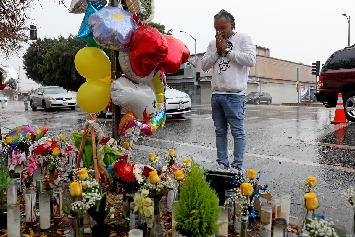A Black man stands with hands to his face, palms together, while he looks at a roadside memorial of candles and balloons. 