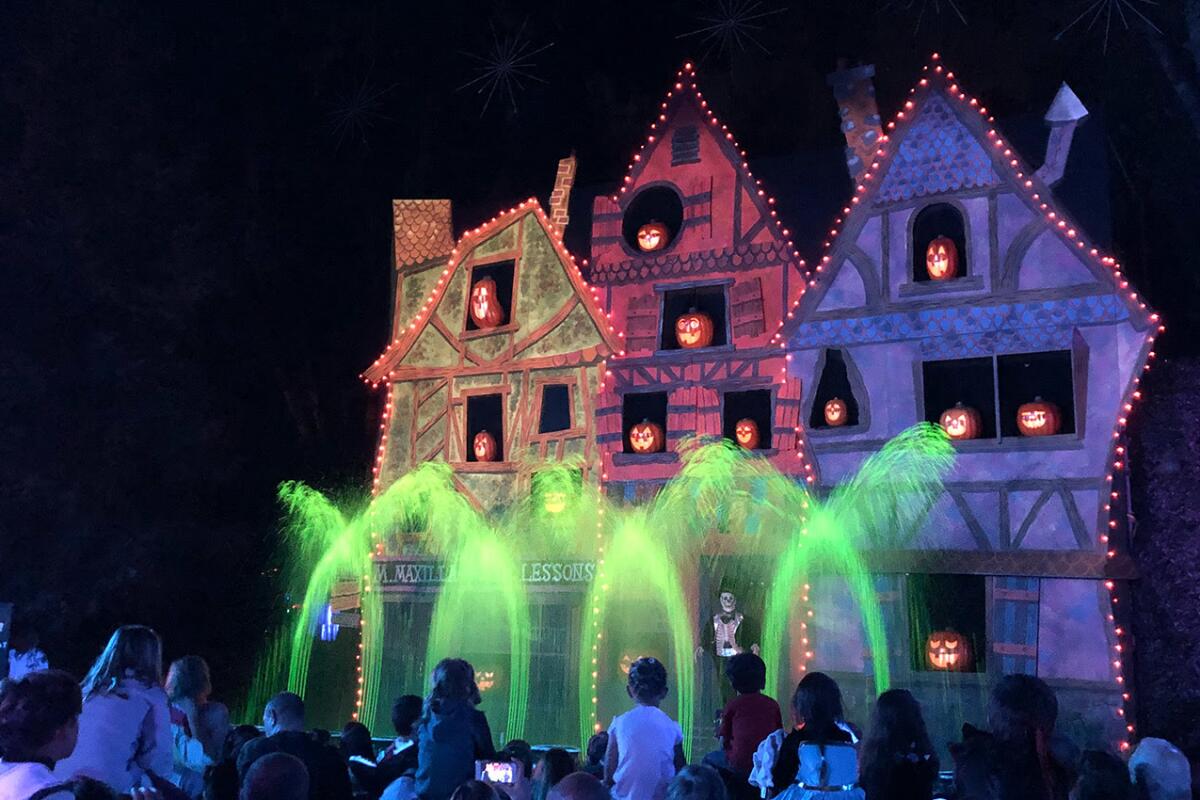People walk in front of colorfully lit scary houses with fountains of green light in front of them