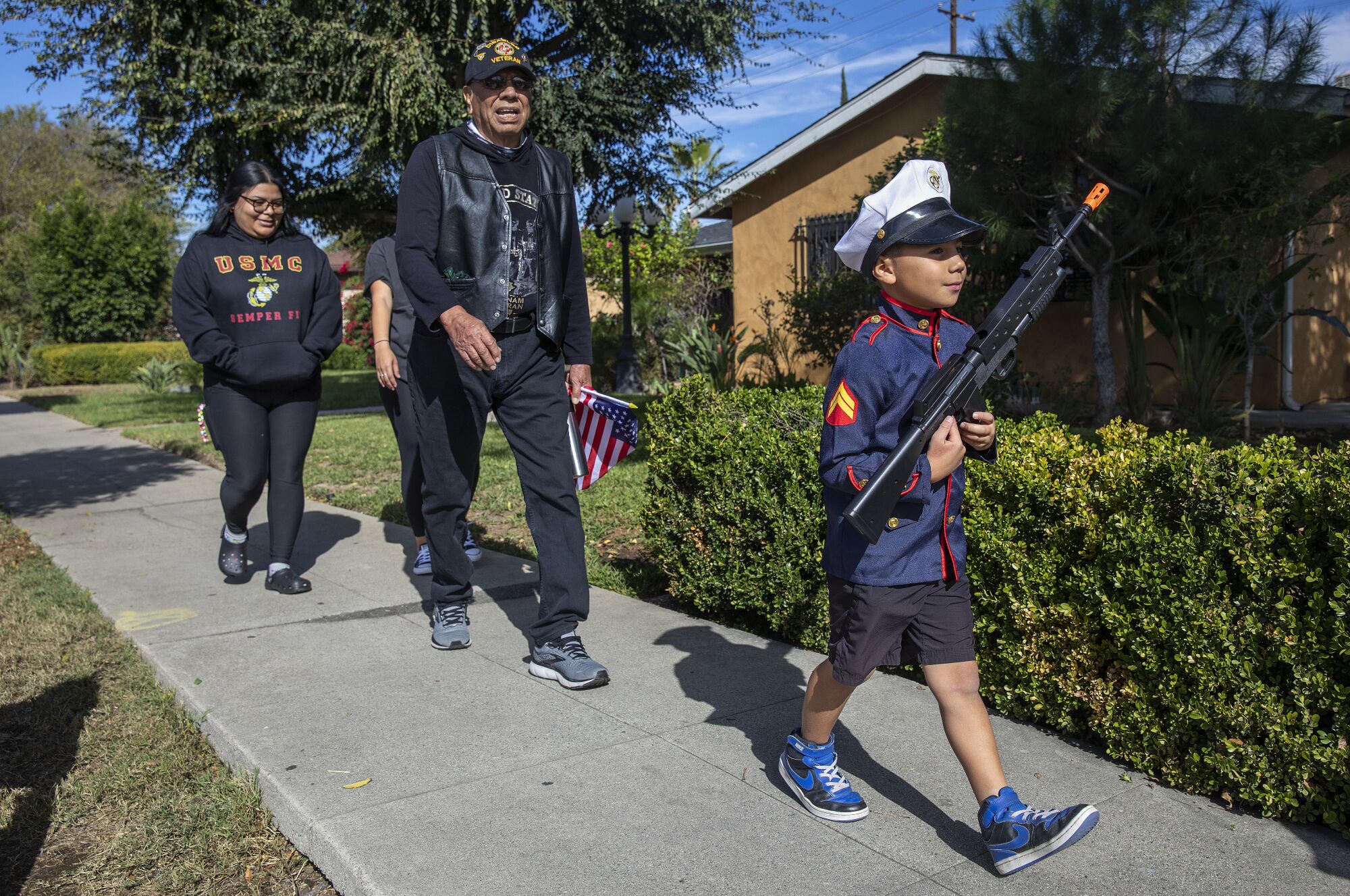 A boy with a toy rifle walks before several adults on a sidewalk 