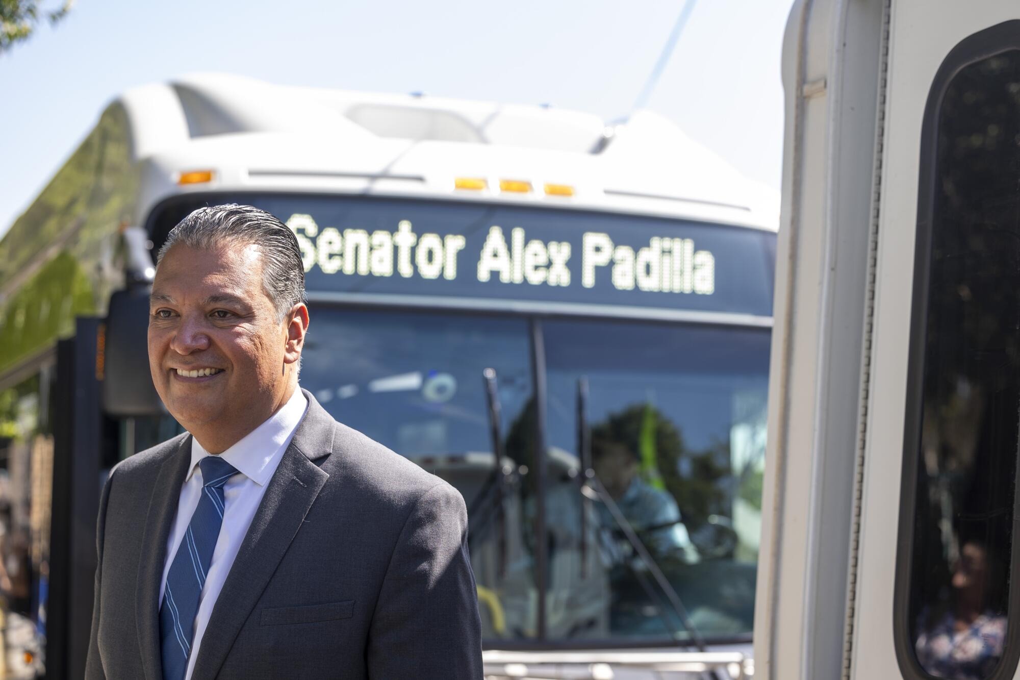 U.S. Sen. Alex Padilla smiles while standing in front of a bus that has his name on the marquee. 