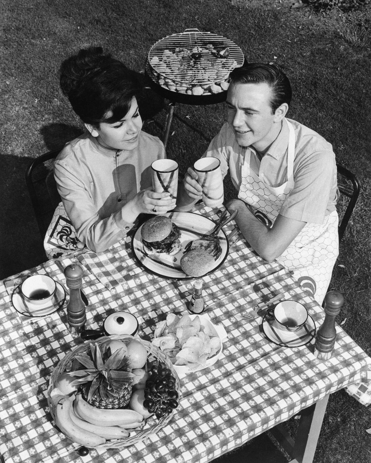 A woman and a man sitting over a meal at a table with a checkered tablecloth, a charcoal grill behind them.