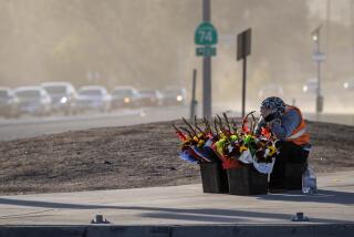 SAN JACINTO, CA - NOVEMBER 24, 2022: Juan Star covers his face as powerful wind gusts blows sand and dirt across the intersection where he is selling flowers on Thanksgiving day on November 24, 2022 in San Jacinto, California. The power is out in the area of Highway 74 and Vista Place. The San Ana winds will continue through Friday.(Gina Ferazzi / Los Angeles Times)