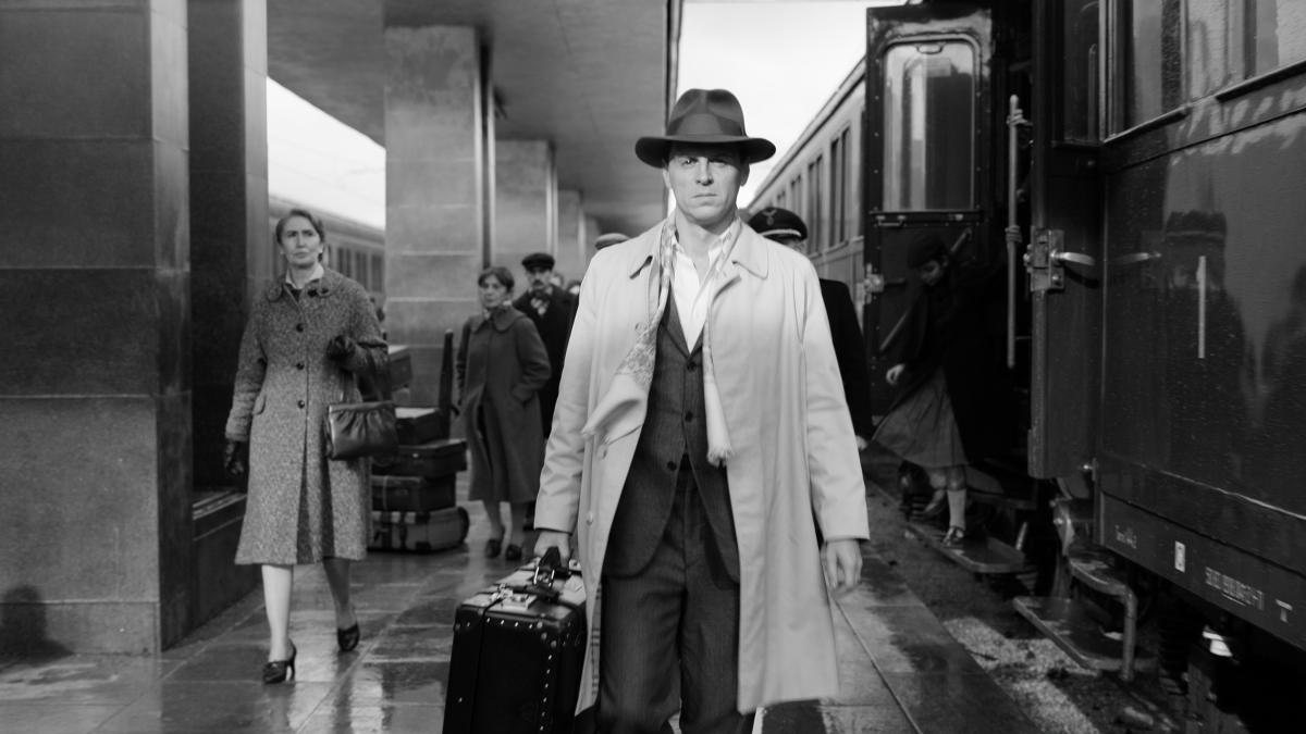A man in a coat and hat carrying a suitcase walking by a train.