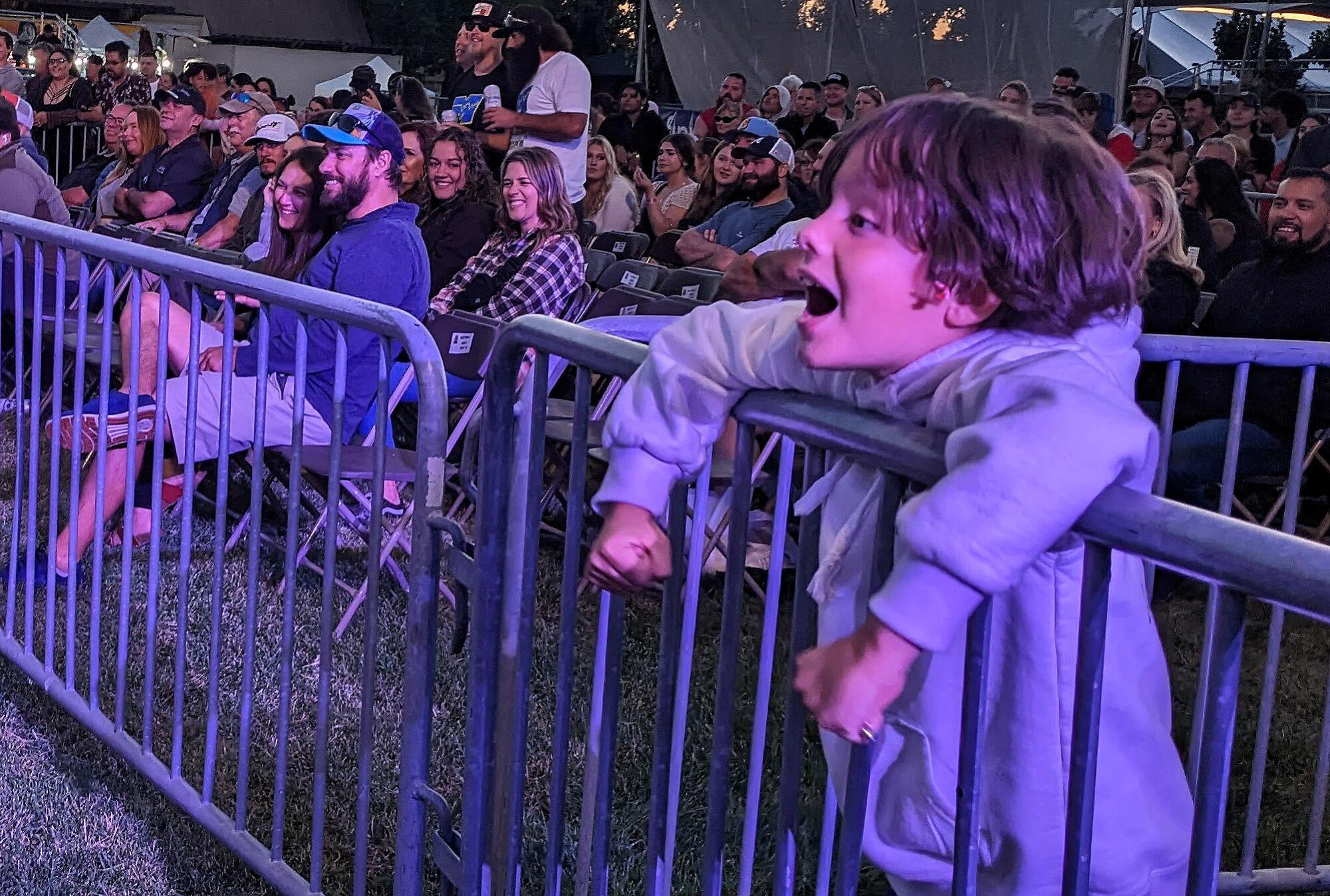 A child behind a metal barricade has his mouth open, with people seated to his right.