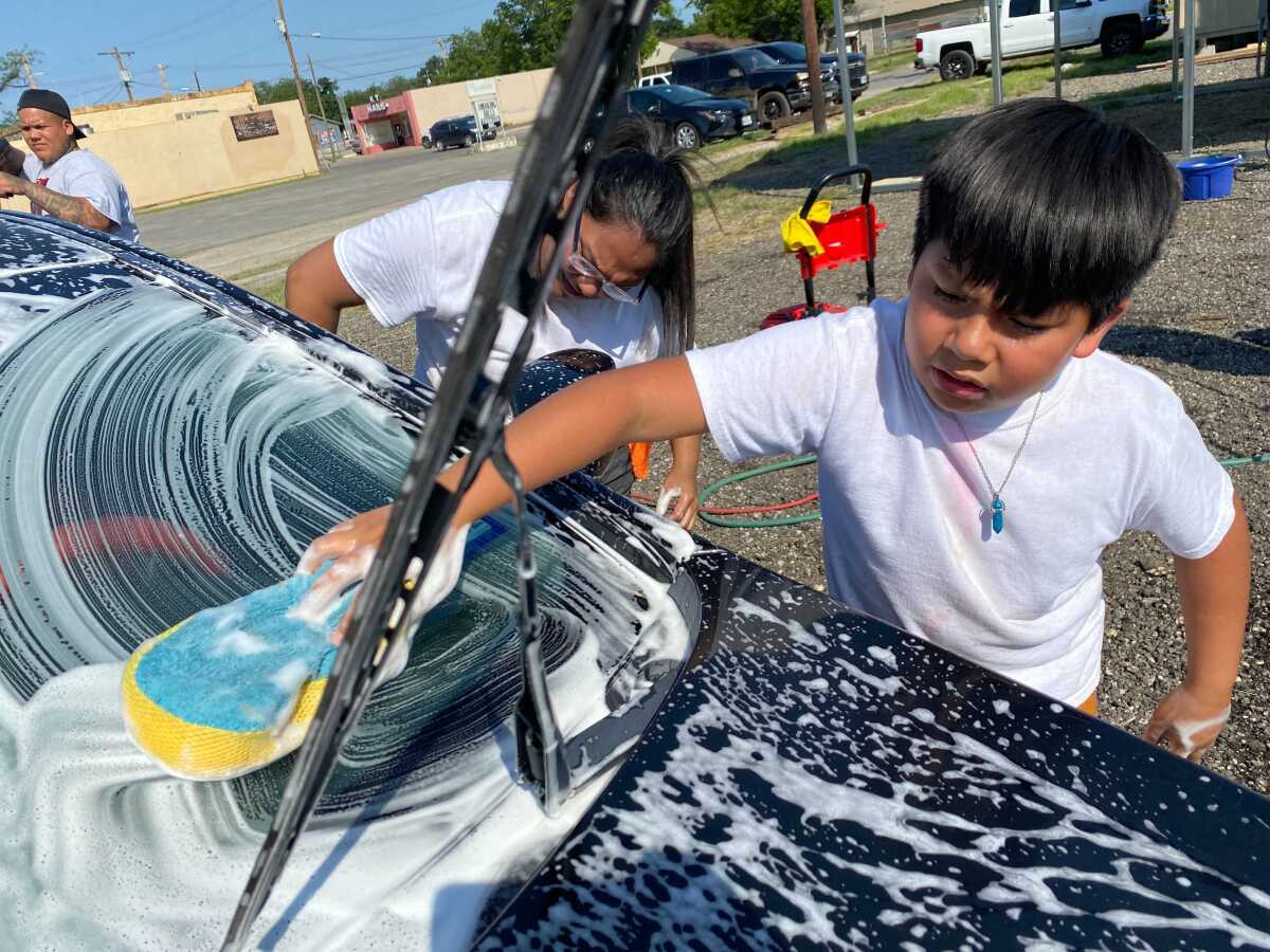 A boy uses a sponge to wash a car windshield covered in soap suds. Next to him is a woman also washing the car