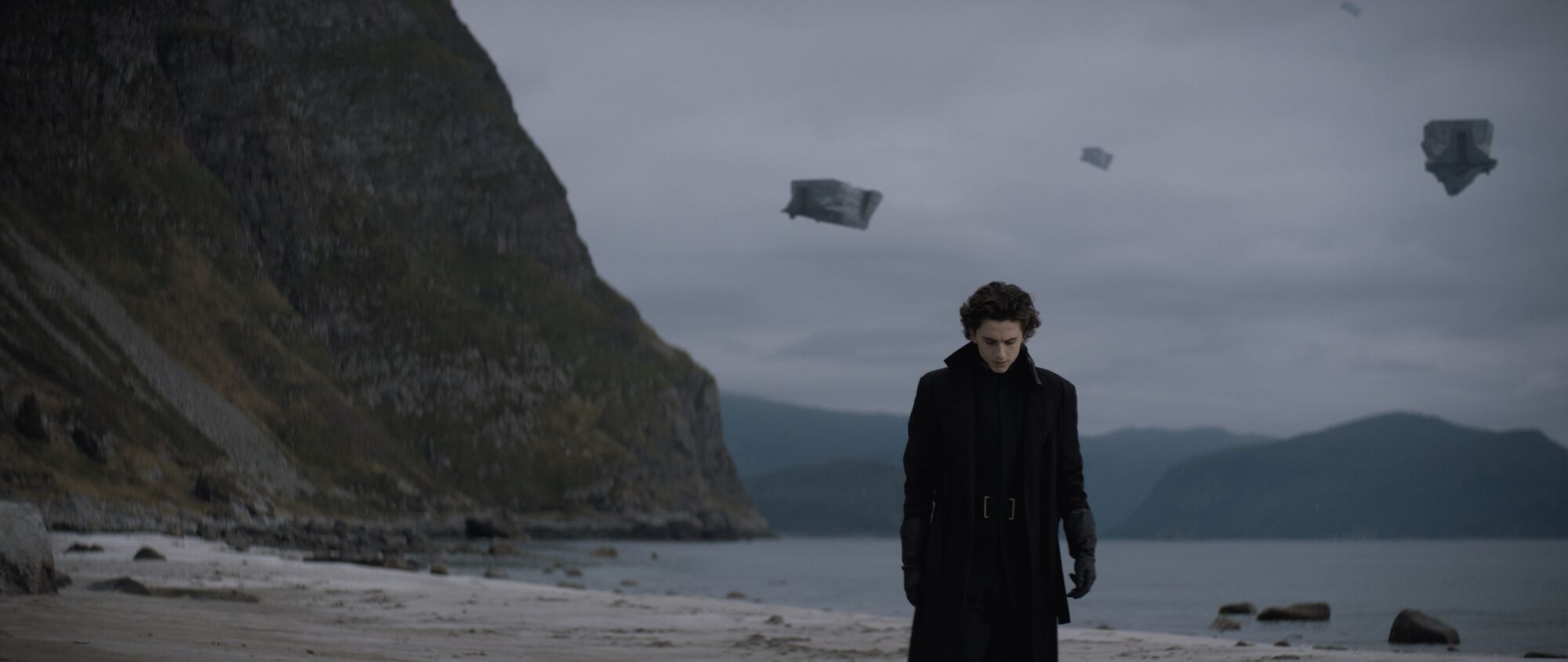 Timothee Chalamet as Paul Atreides walks along the shore as flying ships pass overhead.