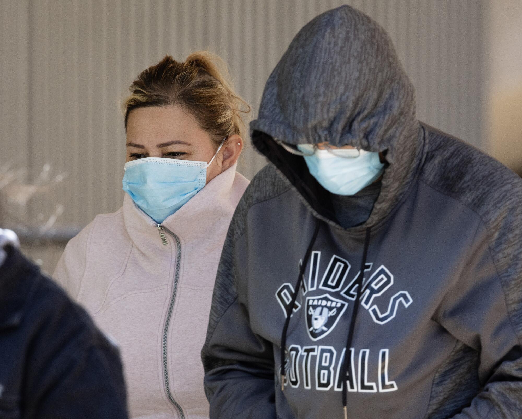 Two people in protective masks walking, one with her collar upturned and one with a Raiders sweatshirt hood over their head