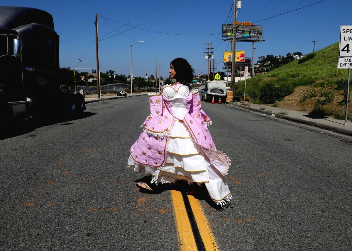 A teen girl walks across a street carrying a pink and white ruffled dress.