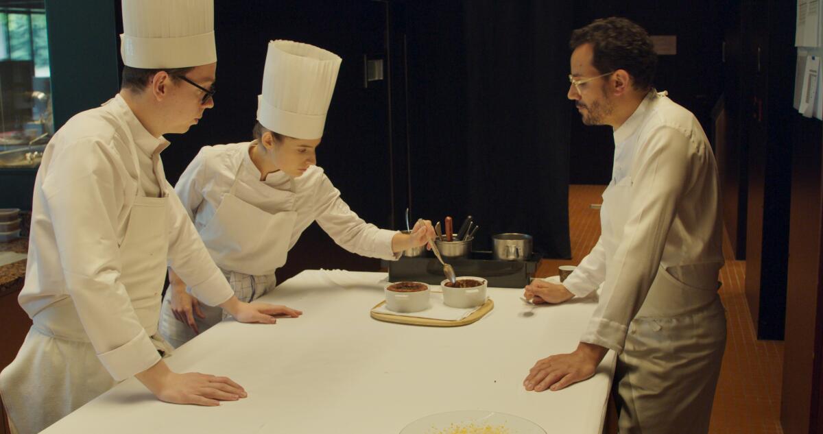 Three French chefs stand at a table, discussing and tasting a dish.