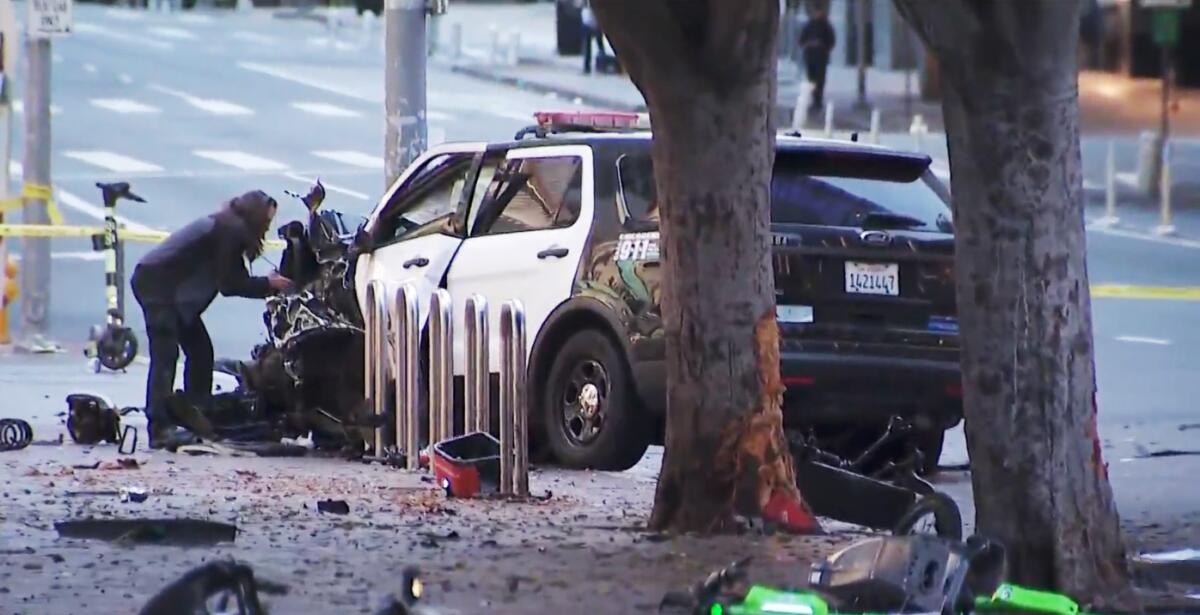 A damaged police vehicle is parked near a lot of debris on the street and sidewalk while a person stands nearby.