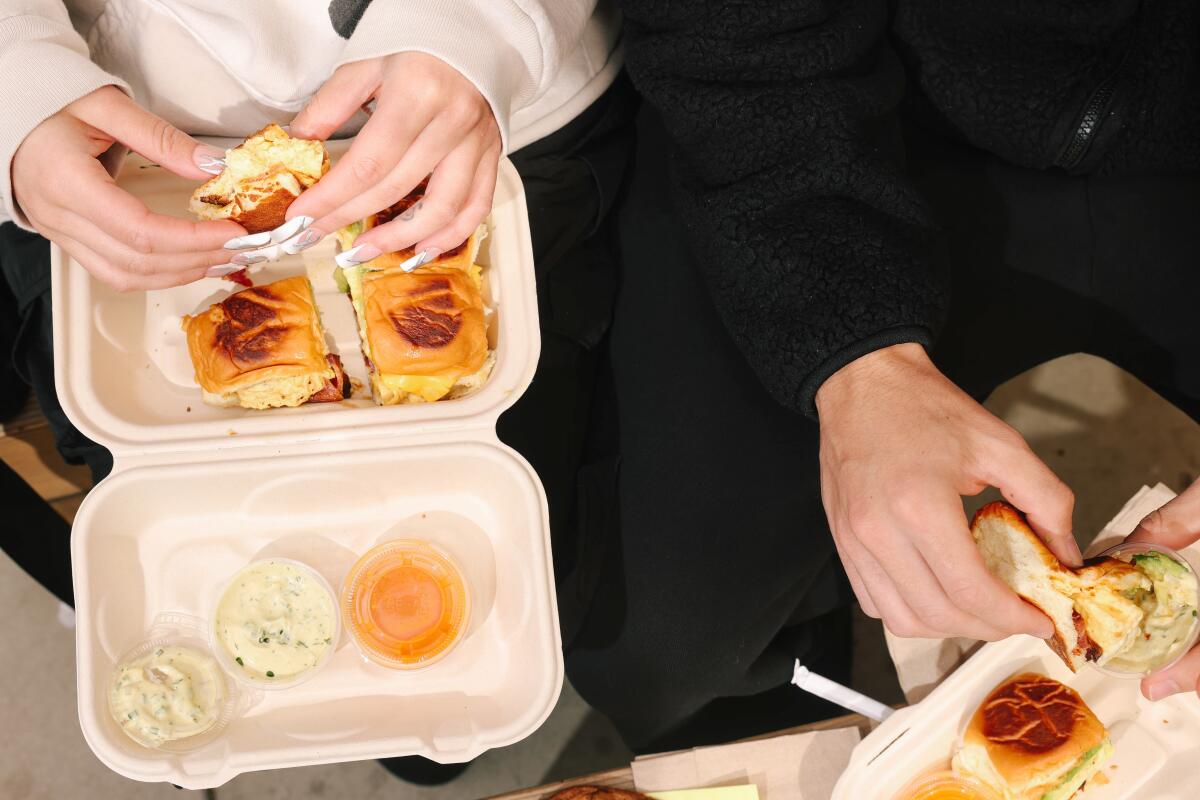 Two people sit close together eating breakfast sliders from to-go containers