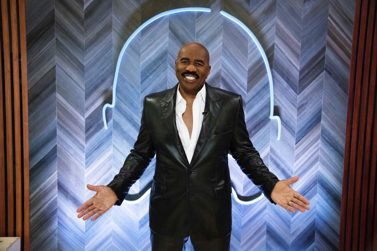 A bald man with a mustache shrugging in a black suit
