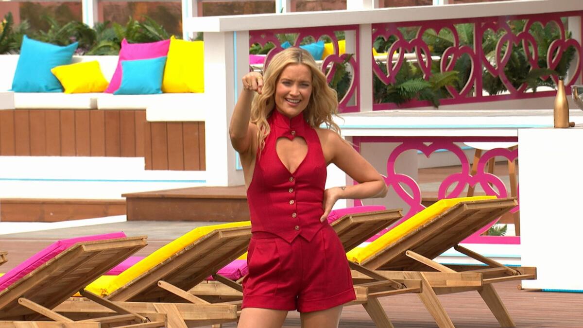 A blond woman in red shorts and top with heart cutout stands next to a row of chaises on a wooden deck.