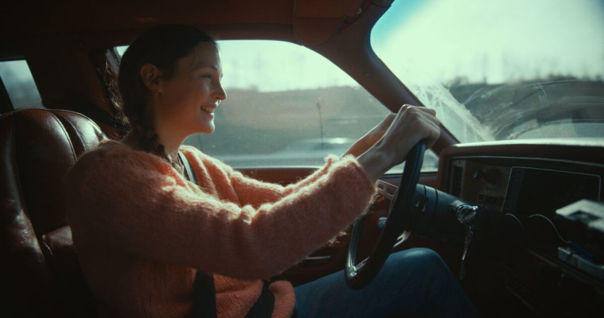A smiling woman in a fuzzy sweater drives a car with a dirty windshield.