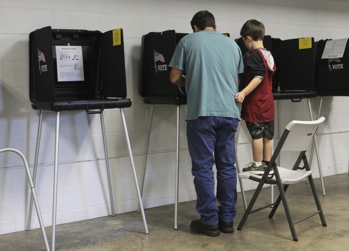 A boy stands on a chair to watch his father at a voting machine.