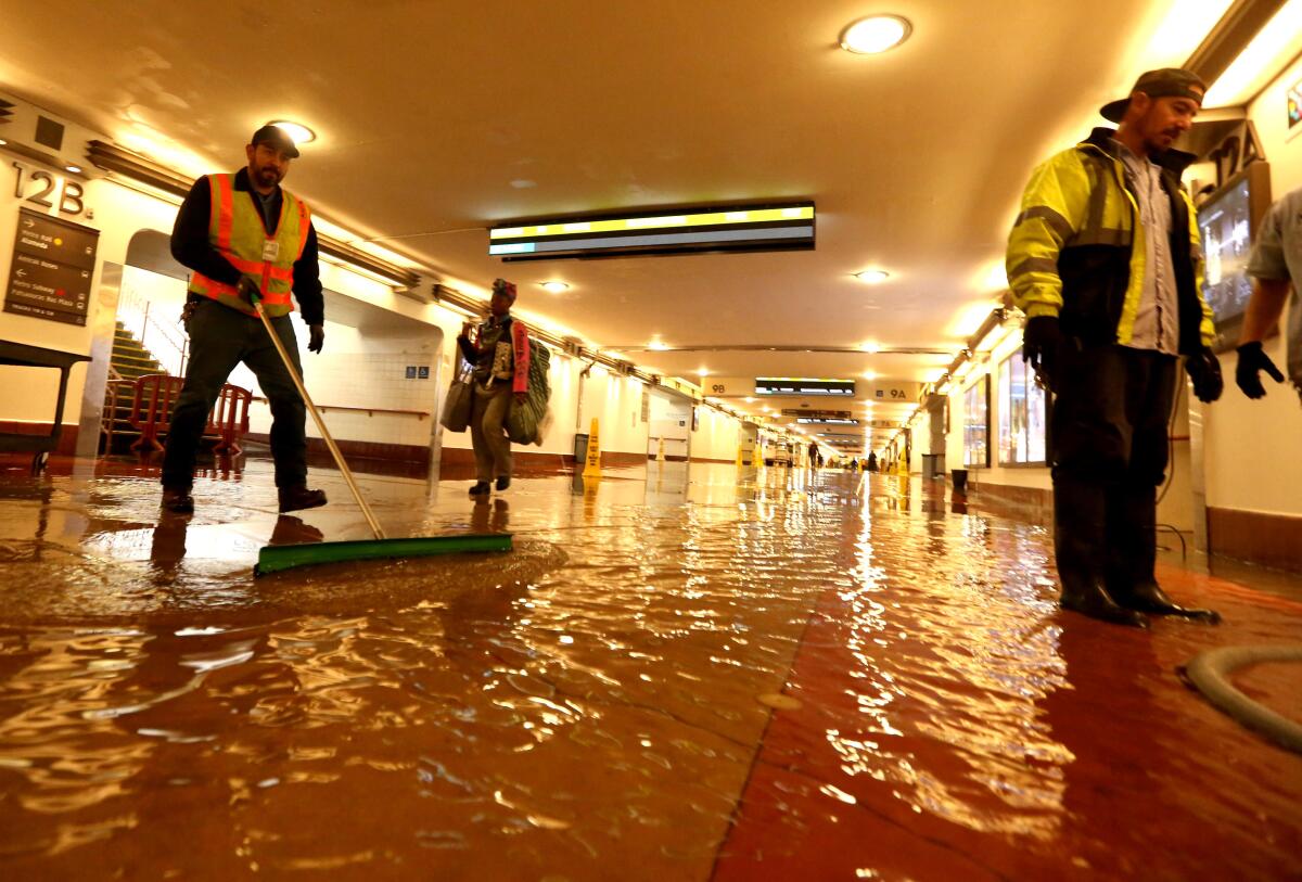 two men in hazard vests try to move water inside a flooded hallway with a red tile floor as a passenger walks through