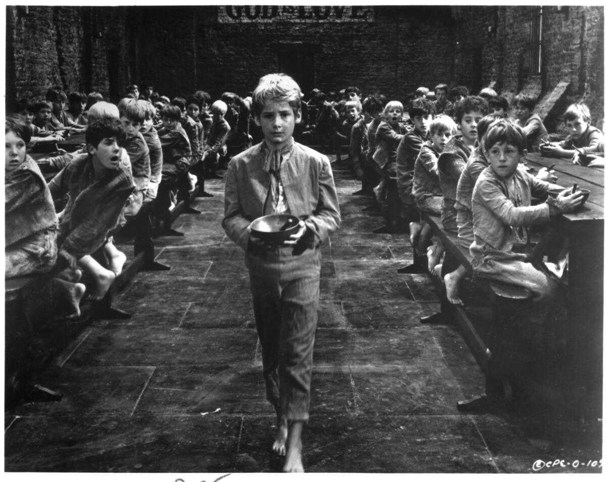 Black and white photo of a young boy walking through a food hall holding a bowl with many boys looking at him