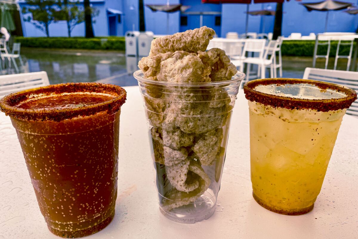 Two drinks in plastic cups with salted rims on either side of a plastic cup full of chicharrones