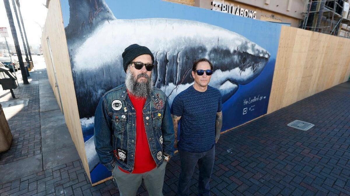 Local artists Hector “HEK” Valdez, left, and Dan McNab stand in front of “The Landlord,” a great white shark mural they painted along Main Street in downtown Huntington Beach.