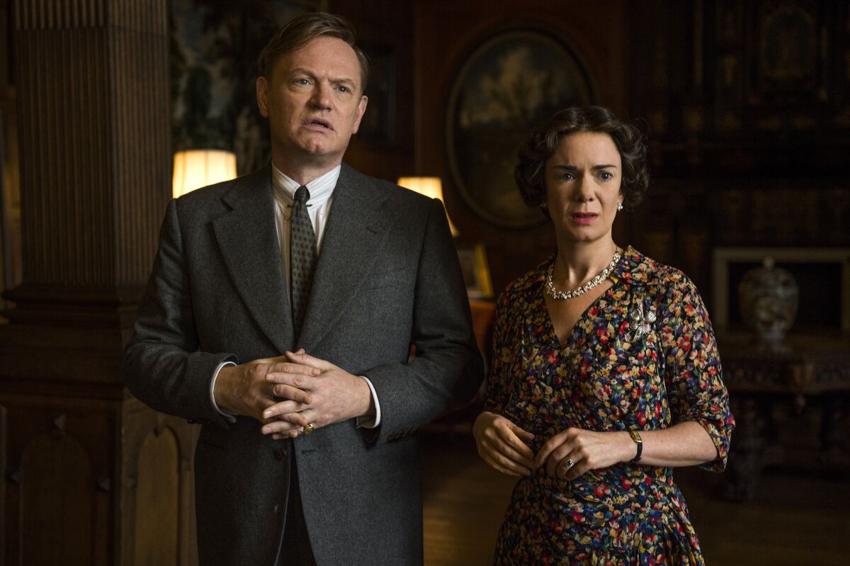 Jared Harris as King George VI in a gray suit standing next to Victoria Hamilton in a floral dress.
