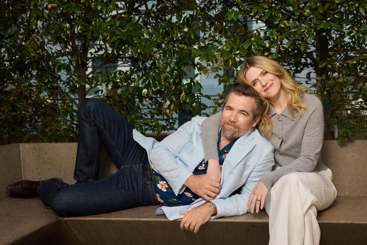 Patrick Brammal and Harriet Dyer huddle on an outdoor bench for a portrait.