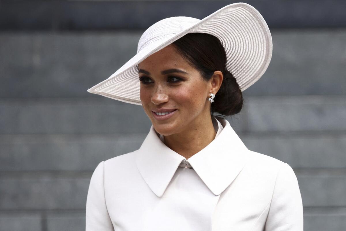 Meghan, Duchess of Sussex, smiles on a staircase while wearing a white coat and hat