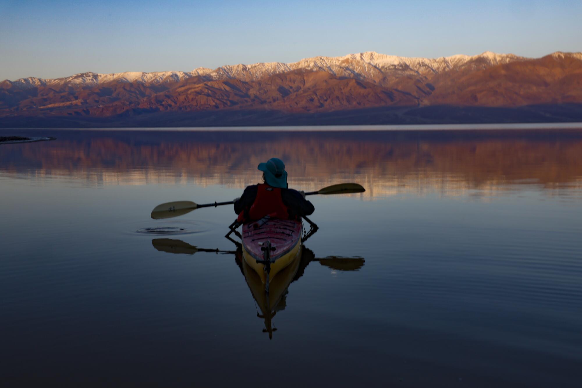 A man in a kayak plies a glassy lake with snowcapped mountains in the distance.