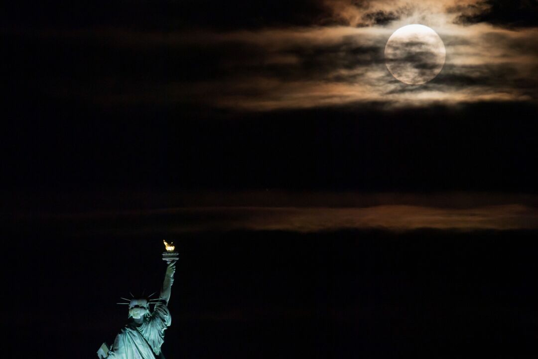 The Statue of Liberty appears to raise her torch toward the rising full moon.
