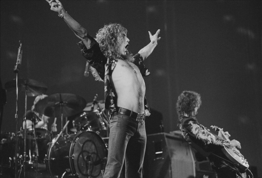 A male rock singer with long curly hair holds his hands up to the sky while performing
