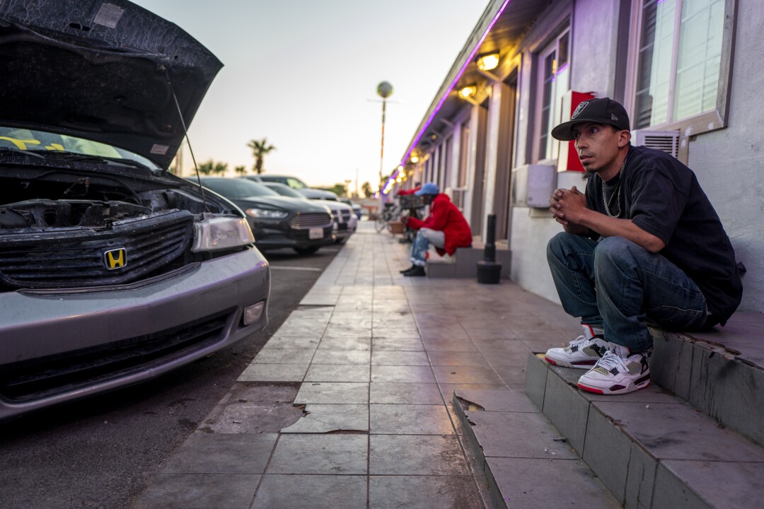 A man in a cap sits on the steps outside a motel room. In front of him is the open hood of a vehicle