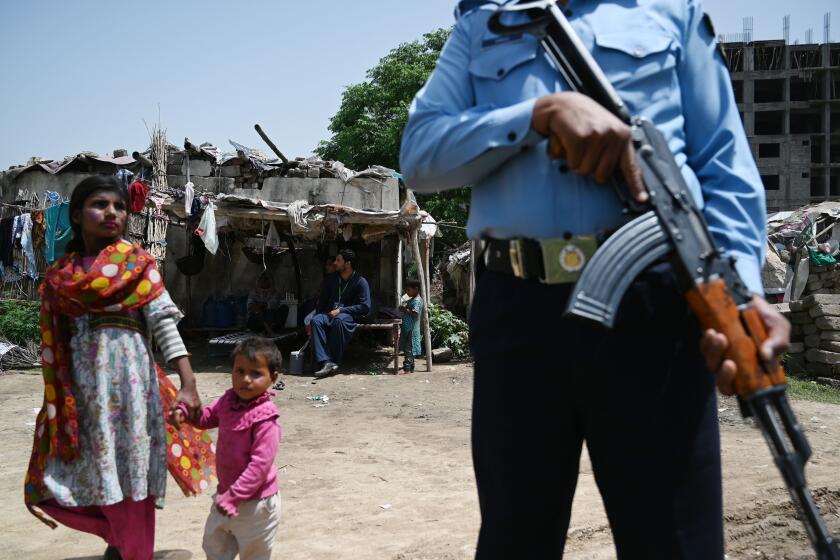 A Pakistani policeman stands guard as health workers in background look on during a door-to-door polio immunization campaign on the outskirts of Islamabad on April 26, 2019. Flanked by armed guards, polio workers fanned out in the slums of Pakistan's capital Islamabad after a bloody week of fatal attacks on health workers threatened to derail an ongoing vaccination drive.