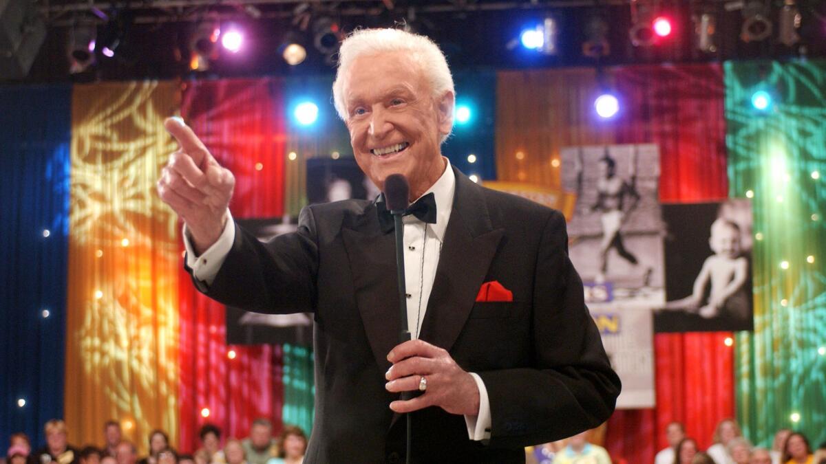 A white-haired man in a tuxedo holds a stick microphone to his mouth and points out of frame in front of a colorful backdrop