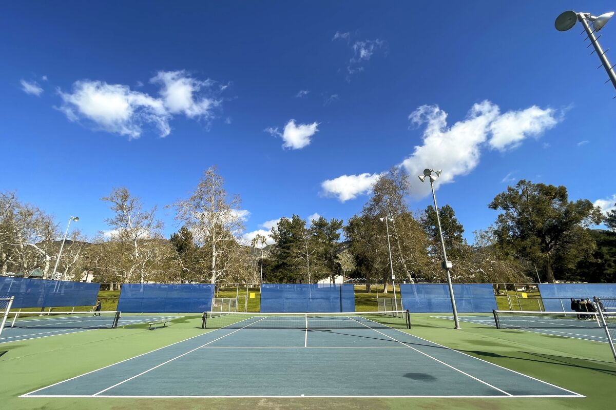 A tennis court with blue backboards in the background, under a deep blue sky with puffy white clouds. 