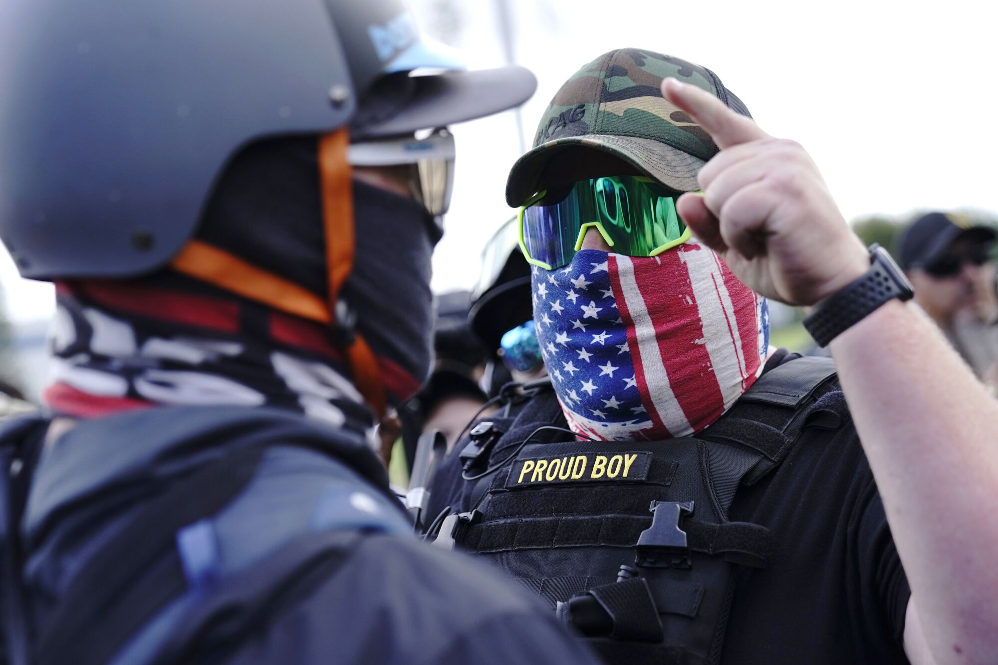 A man in a U.S. flag mask and tactical vest with a Proud Boy patch points into the masked face of a counter-protester