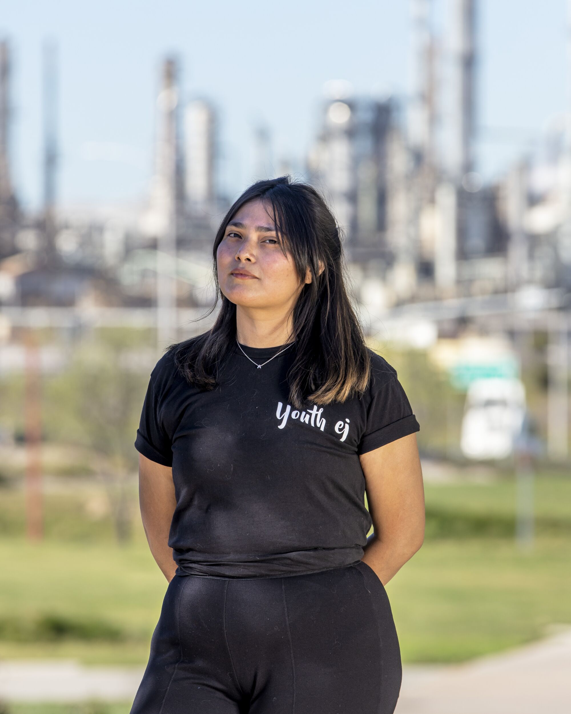 A young woman stands in front of an oil refinery