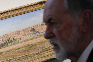 A man with a gray beard next to a painting of a desert landscape with buildings and trees