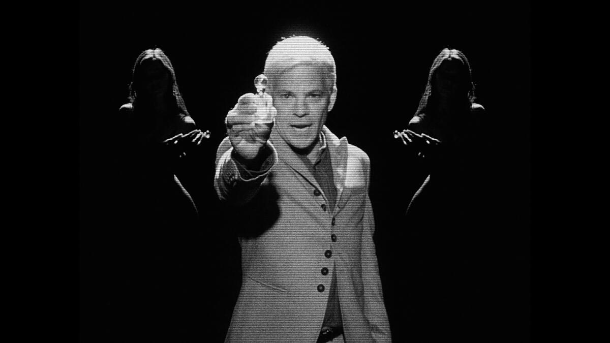 In a black-and-white film still, a man holds up a product he's selling. Two women stand in the shadows behind him.