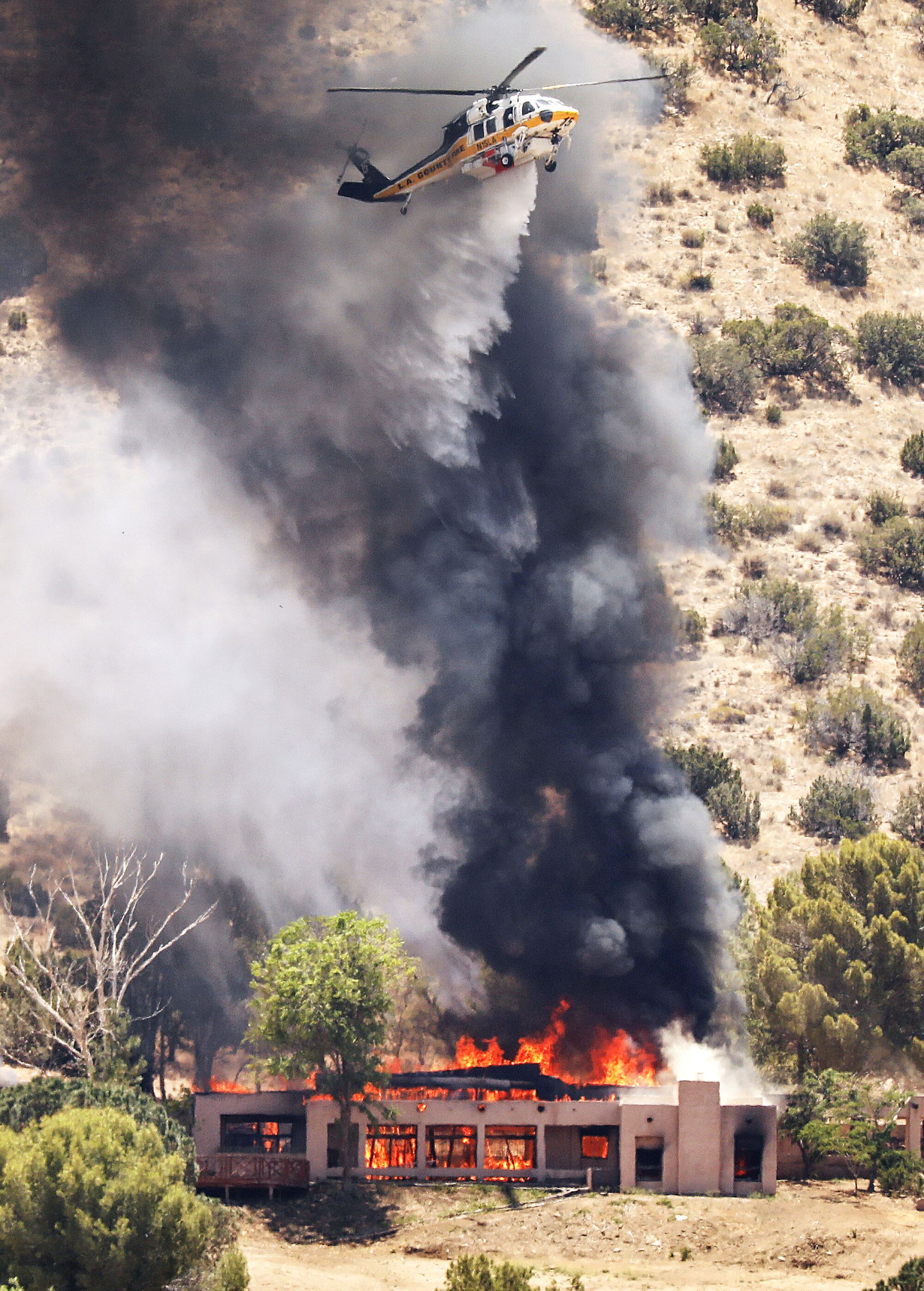 A  helicopter hovers amid a black plume of smoke as a house below is fully engulfed in flames.