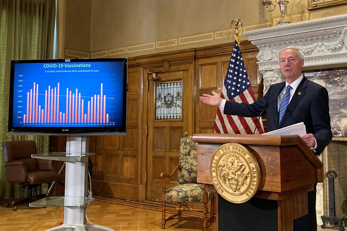 Asa Hutchinson, behind a lectern that bears the state seal, gestures to a screen labeled "COVID-19 vaccinations." 