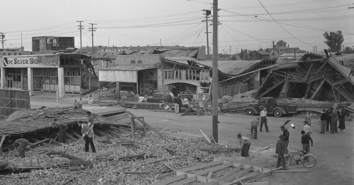 People walk along a road next to heavily damaged shops.